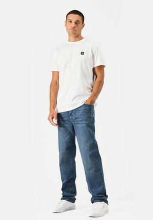 Junger Mann steht, trägt ein weißes kurzärmliges T-Shirt, blaue Jeans und weiße Sneakers vor einem schlichten hellen Hintergrund.