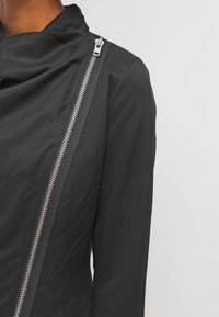Chaqueta negra con un detalle de cremallera plateada en diagonal. Tejido suave, corte entallado y diseño de cuello drapeado. Estilo minimalista.