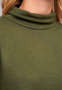 Haut vert olive avec un col roulé haut. Fabriqué en tissu doux et extensible avec une texture lisse, présentant un design minimaliste et sans motifs.