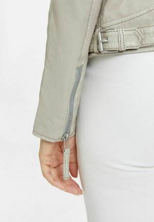 Manche de veste en cuir gris clair avec une fermeture éclair argentée et un détail de sangle. Présente une texture lisse et une couture contrastante près du poignet.