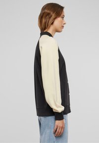 Kvinne i svart og krem varsity-jakke med lyseblå jeans, stående i profil mot en enkel grå bakgrunn.
