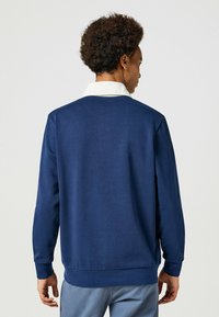 Granatowy sweatshirt z białym kołnierzykiem, wykonany z miękkiego materiału, cechujący się prostym wzornictwem i ściągaczami na rękawach. Widok z tyłu pokazuje gładką fakturę.