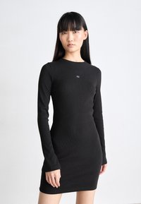 Black long-sleeve ribbed dress with a round neckline and a small logo on the chest. The fabric has a smooth texture and fitted silhouette.