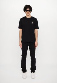 Juodas medvilnės marškinėliai su mažą auksinę logotipą ant krūtinės, derinami su juodomis džinsomis ir baltais sportbačiais, modelis dėvi tamsius akinius nuo saulės.