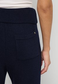 Gebreide marineblauwe broek heeft een brede ribgebreide tailleband, achterzakken en een gouden accentdetail. De textuur lijkt zacht en comfortabel.