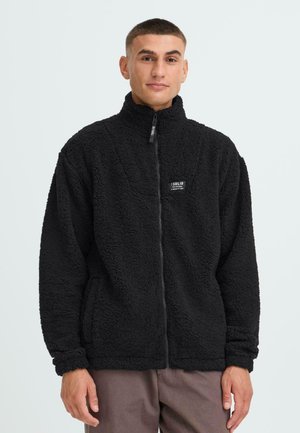 Schwarze Fleecejacke mit hohem Kragen und durchgehendem Reißverschluss, mit strukturiertem Material und einem kleinen Logopatch auf der linken Seite.
