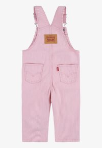 Salopette en denim rayée rose et blanche avec des bretelles ajustables, poche avant et poches arrière plaquées. Étiquette de marque en cuir marron sur la poitrine.