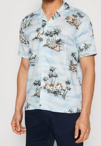 Chemise à manches courtes boutonnée en tissu bleu clair avec un imprimé tropical comportant des palmiers, des huttes et des planches de surf, texturée avec une finition lisse.