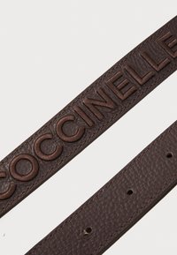 Cinto de couro castanho com nome da marca em relevo, superfície texturizada e vários furos para ajuste de comprimento.