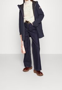 Femme portant une veste bleu marine, un pull à col roulé crème, un jean large foncé, des chaussures à plateforme marron, et tenant un sac à main rose.