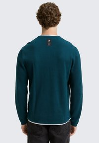 Türkiser Pullover mit langen Ärmeln und gerippter Textur sowie Rundhalsausschnitt. Mit einem schwarzen Stofflabel auf der Rückseite, kombiniert mit dunklen Jeans.
