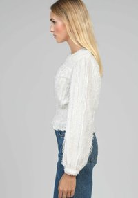 Femme aux longs cheveux blonds portant un blouse blanche à manches longues texturée et un jean bleu, se tenant de côté devant un fond gris.