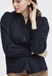 Námornícka blúzka s légiou a gombíkovým zapínaním, s nariasenými detailmi na výstrihu a rukávoch, a so zosilnenými rukávmi.