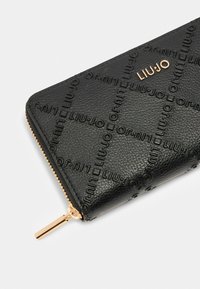 Portefeuille en cuir texturé noir avec un design de logo embossé, des accessoires zippés en ton doré et une forme rectangulaire. Compact et fonctionnel.