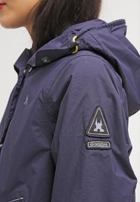 Navy jacket with a hood, featuring a textured fabric, zippered closure, and a logo patch on the sleeve. Accented with yellow cord.