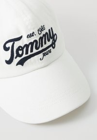 Casquette de baseball blanche en coton avec le texte "Tommy Jeans" brodé en bleu marine. Présente une couronne arrondie et un bord incurvé. Design simple et épuré.