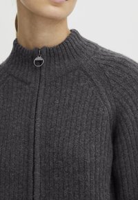 Ingrandimento di una persona che indossa un maglione zip-up a coste grigio scuro con colletto alto e zip circolare.