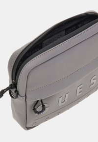 Bolso bandolera sintético gris con un acabado texturizado. Presenta un logo embossado, cierre con cremallera y costuras sutilmente contrastantes.