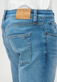 Világoskék farmernadrág bőr címkével, amelyen a "TOMMY JEANS" felirat olvasható, a derékszíjnál. Két hátsó zsebbel rendelkezik, kis logós részletekkel.