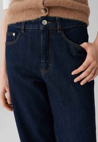 Donkerblauwe denim jeans hebben een hoge taille, zijzakken en contrasterende stiksels. De stof ziet eruit als soepel met een lichte glans.