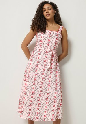 Vrouw met krullend haar, gekleed in een mouwloze witte jurk met rode bloemen- en geometrische patronen, staand met de handen achter haar rug.