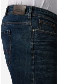 Jeans in denim blu scuro con una toppa in pelle etichettata "JOHN E. GEE." Presentano cuciture visibili e una tasca laterale con un rivetto.