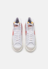 Valkoiset nahkaiset korkeat Nike urheilukengät, joissa on harmaat mokkanahkaiset yksityiskohdat, oranssi swoosh ja keltaiset kielivärit. Varustettu valkoisilla nauhoilla.