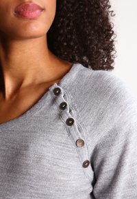 Szary sweter z dekoltem w serek, z czterema ciemnymi guzikami na lewym ramieniu oraz miękką, teksturowaną tkaniną.