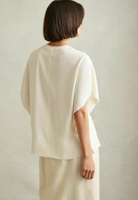 Top color crema a maniche corte con vestibilità rilassata, caratterizzato da una cucitura lungo il retro e un tessuto fluido che cade morbido.