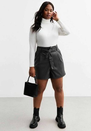 Shorts en simili cuir noir avec une taille ceinturée, associés à un col roulé blanc et des bottines noires, tenant un petit sac à main noir.