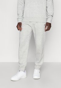 Γκρι φόρμα από απαλό ύφασμα με ελαστικά μανσέτες, με λογότυπο ραμμένο στο αριστερό μηρό και συνδυασμένη με λευκά αθλητικά παπούτσια.