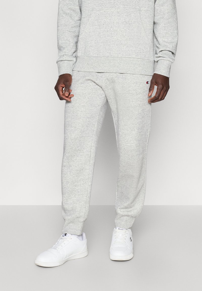 Γκρι φόρμα από απαλό ύφασμα με ελαστικά μανσέτες, με λογότυπο ραμμένο στο αριστερό μηρό και συνδυασμένη με λευκά αθλητικά παπούτσια.