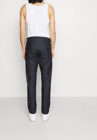 Dark blue denim jeans with standard pockets, subtle stitching details, and a straight-leg cut, worn with a white tank top and white sneakers.