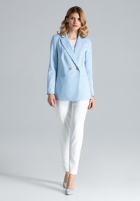 Jasnoniebieska marynarka dwurządowa wykonana z gładkiego materiału z złotymi guzikami, zestawiona z białymi spodniami i srebrnymi szpilkami, stojąca na neutralnym tle.