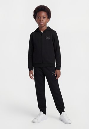 Αγόρι που φοράει μαύρο φούτερ με φερμουάρ και αντίστοιχο παντελόνι φόρμας με λευκά αθλητικά παπούτσια, στέκεται μπροστά σε απλό λευκό φόντο.