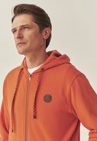 Oranje hoodie met ritssluiting, gemaakt van zachte stof, met een ronde bruine patch met een berenlogo, zwarte gestreepte trekkoorden en een klassieke pasvorm.