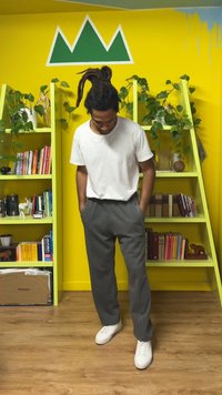 Pantalon gris à coupe décontractée, associé à une chemise blanche à manches courtes et des baskets blanches. En arrière-plan, un mur jaune vif et des étagères.