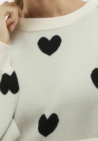 Suéter de color crema con patrones de corazones negros, tejido texturizado, escote amplio y puños de canalé. Vista en primer plano del detalle de la tela.