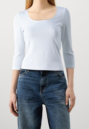 Woman wearing a plain white three-quarter sleeve top and blue high-waisted jeans, standing against a neutral background.