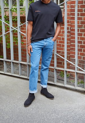 Mand iført sort kortærmet t-shirt, lyseblå opsmøgede jeans, sorte ruskindsstøvler, stående med den ene hånd i lommen ved et metalhegn og en murstensvæg.