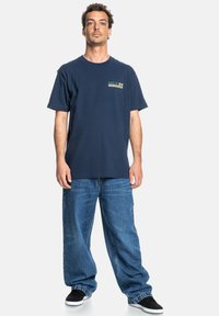 Navy blue cotton t-shirt with short sleeves and a logo in green and yellow. Paired with loose-fitting blue denim jeans and black slip-on shoes.