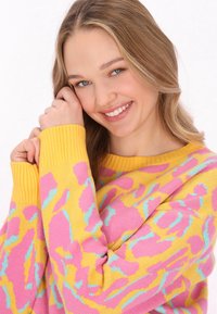 Barevný pletený svetr s žlutým základem a abstraktními vzory v růžové a tyrkysové barvě, s žebrovanými manžetami a volným střihem.