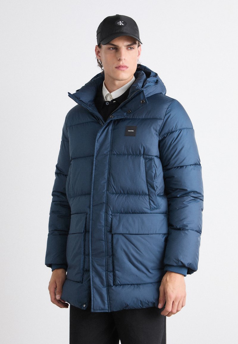 Parka imbottita blu navy con cappuccio, zip anteriore, bottoni a pressione, due grandi tasche anteriori e un piccolo logo sul petto.