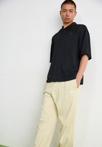 Μαύρο πόλο μπλουζάκι με κοντά μανίκια που διαθέτει λογότυπο στο στήθος, σε συνδυασμό με ανοιχτό μπεζ φόρμες που έχουν λογότυπο στο πλάι.