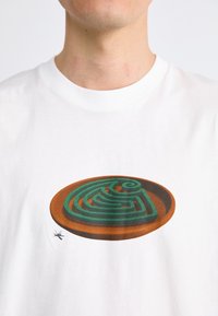 Hvit t-skjorte med et brunt sirkulært maze-design med grønne konturer, sammen med en liten insektgrafikk nær bunnen.