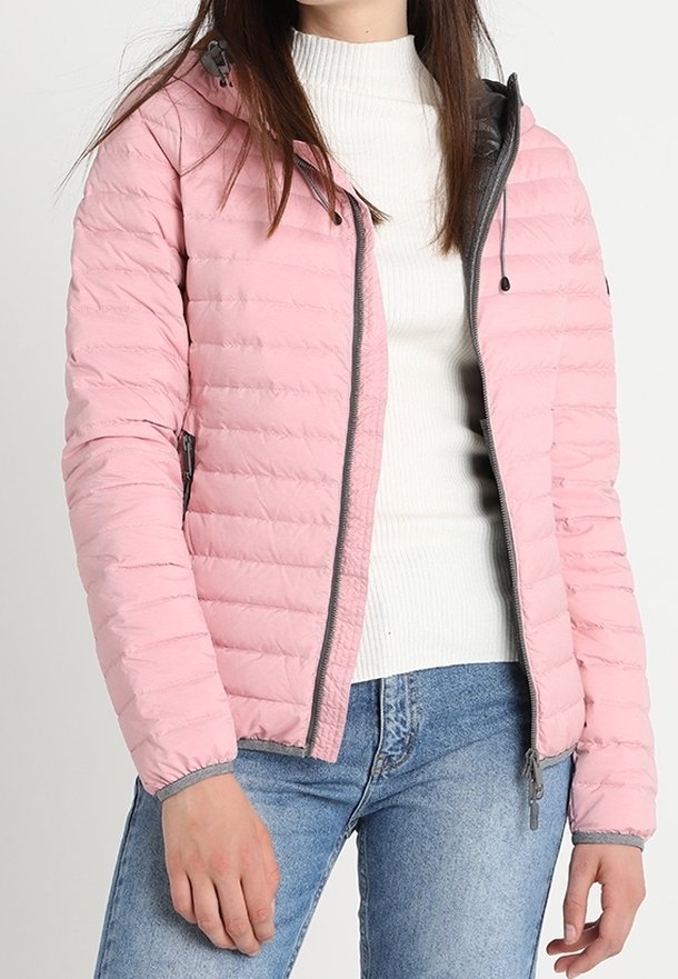 Femme portant une veste matelassée rose clair par-dessus un pull à col roulé blanc côtelé et un jean en denim bleu.