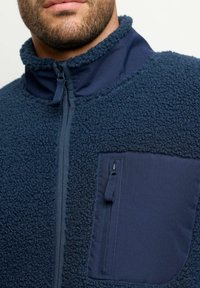 Giacca in pile blu navy con colletto alto e zip frontale. Presenta una tasca in tessuto liscio sul lato sinistro del petto con chiusura a zip. Superficie spessa e strutturata.