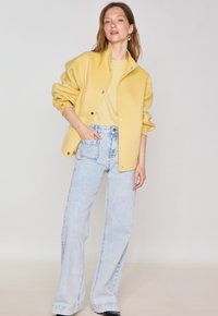 Yellow oversized jacket with snap closures, paired with a light yellow top and high-waisted light blue flared jeans. Smooth texture throughout.