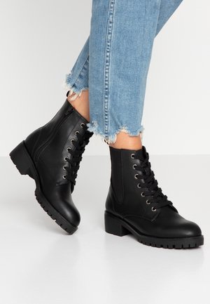 Pieds chaussés de bottines noires en cuir à lacets avec des fermetures éclair latérales, associés à un jean bleu clair à ourlet effiloché, sur fond blanc.