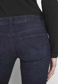 Jeans de denim azul oscuro con una textura suave, que presentan dos bolsillos traseros y un parche de cuero negro con la marca en la cinturilla.